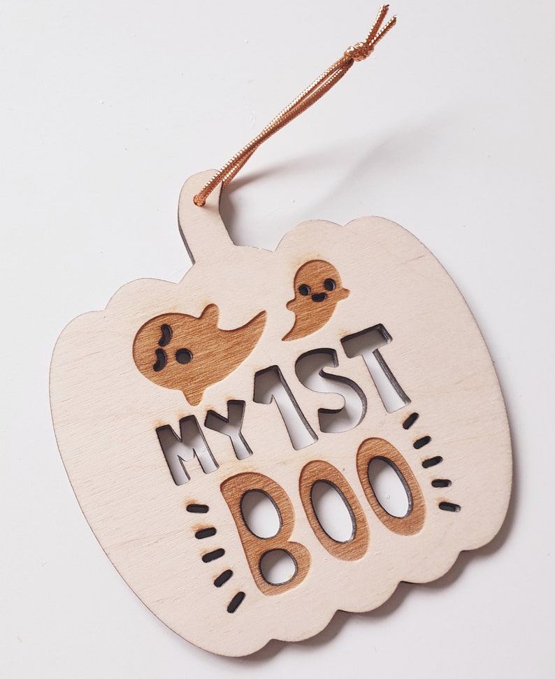 May include: A wooden pumpkin-shaped ornament with the text "MY 1ST BOO" and two ghost cutouts. The ornament is painted in a light wood color and has a brown string for hanging.