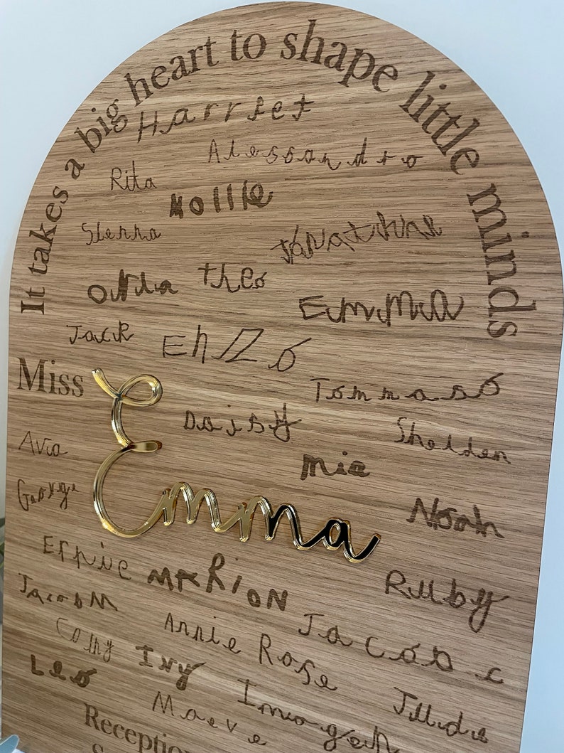 May include: A wooden plaque with the text "It takes a big heart to shape little minds."  The plaque is decorated with a gold-colored name "Emma" and many other names written in cursive.