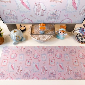 May include: A rectangular desk mat featuring a repeating pattern of pink and blue illustrations, including castles, dresses, and character figures in frames. The mat is positioned on a white surface in front of a computer monitor with a matching wallpaper.