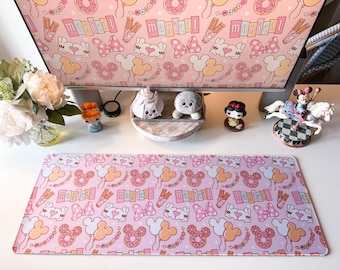 May include: A rectangular desk mat featuring a repeating pattern of pink and light purple backgrounds with illustrations of donuts, bows, churros, and the word magical in rectangular boxes. The design includes cartoon hands forming heart shapes.