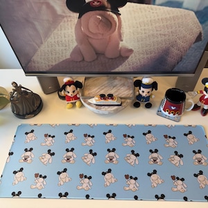 May include: A rectangular desk mat featuring a repeating pattern of light pink plush rabbit figures wearing black mouse-ear hats on a light blue background. The mat has a smooth surface and rounded corners, displaying a consistent grid of the illustrated characters.