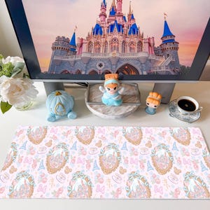 May include: A rectangular desk mat featuring a repeating pattern of gold-framed mirrors, blue wizard hats, pink bows, and teacups on a light pink background. The mat is positioned on a white surface in front of a computer monitor displaying a castle.