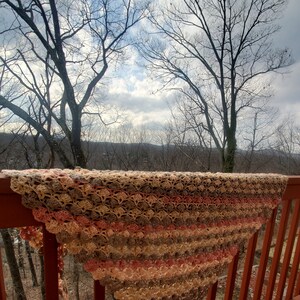 May include: A crocheted shawl in shades of brown, beige, and pink, draped over a wooden railing with a view of a forest in the background.