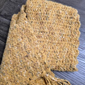 May include: Mustard yellow scarf with a textured, speckled knit design. The scarf has a fringed edge and is folded. The color is a warm, earthy tone, perfect for fall or winter wear. The scarf is made of a soft, cozy material.