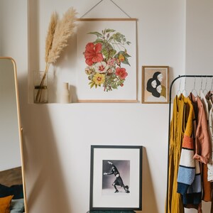 May include: A framed botanical print featuring a vibrant arrangement of red, pink, and yellow hibiscus flowers. The print is displayed with a wooden frame and a metal hanger. Other framed art and clothing are also visible.