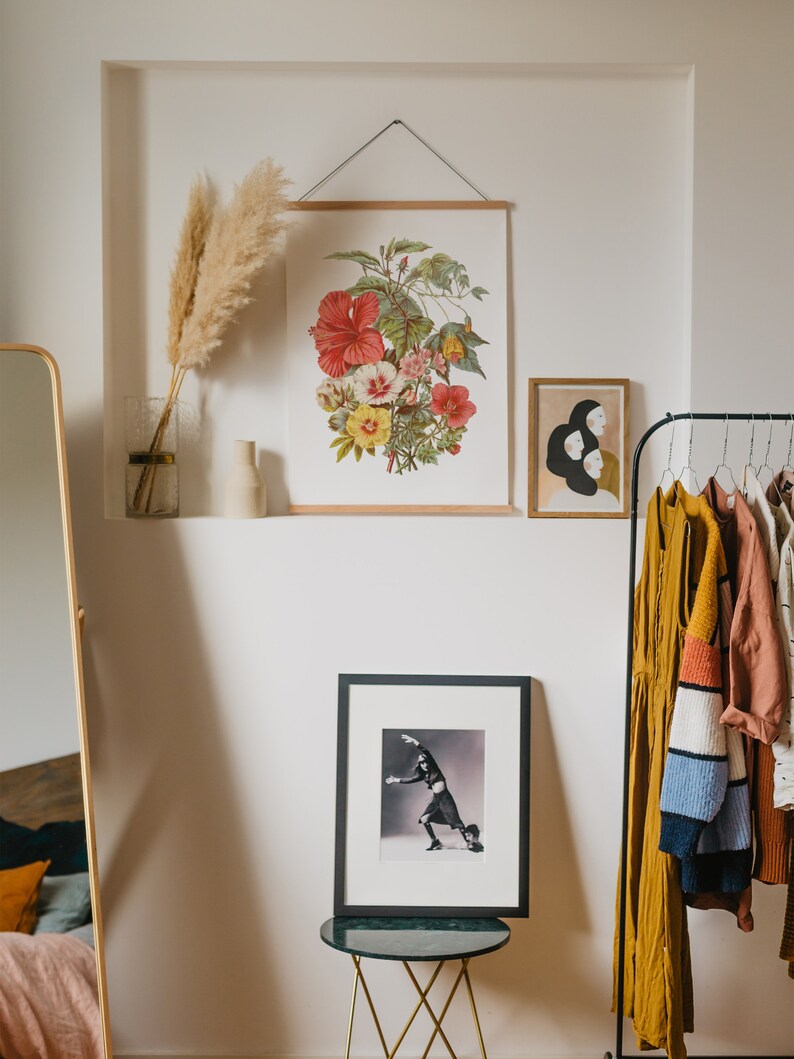 May include: A framed botanical print featuring a vibrant arrangement of red, pink, and yellow hibiscus flowers. The print is displayed with a wooden frame and a metal hanger. Other framed art and clothing are also visible.