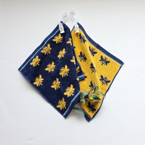 May include: Two square terry towelling cloths featuring a bee pattern. One cloth is navy blue with yellow bees, and the other is yellow with navy blue bees. Each cloth has a white fabric hanging loop attached with a small white button at the corner.