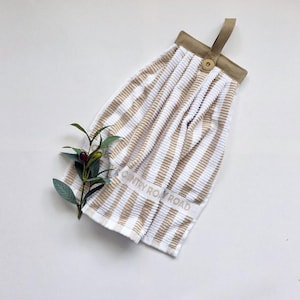 May include: A striped kitchen towel in light brown and white, with the text "COUNTRY ROAD". It features a hanging loop and button detail. A sprig of olives is placed near the towel. The towel is designed for the kitchen.
