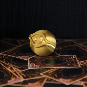 May include: A gold-colored metal object with a raised eye design, resembling a playing card, resting on a surface of black playing cards.