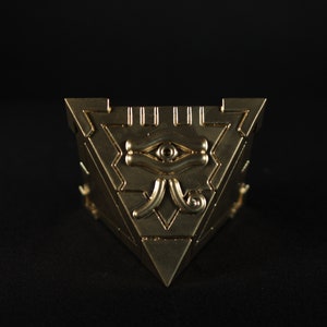 May include: A gold-colored triangular box with an intricate design featuring an eye symbol. The box is a replica of the Millennium Puzzle from the popular anime and manga series Yu-Gi-Oh!