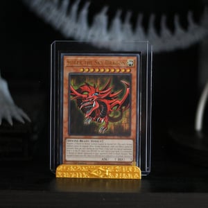 Puede incluir: Una carta coleccionable de Yu-Gi-Oh!, "Slifer the Sky Dragon", en una funda protectora. La carta presenta una ilustración de dragón rojo con texto. La carta se muestra en un soporte dorado.