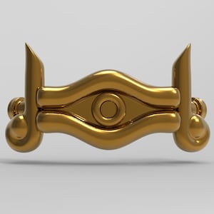 May include: A gold-coloured, three-dimensional replica of the Eye of Horus, a symbol from ancient Egyptian mythology. The eye is depicted with a central circle and a surrounding ring, all within a stylized, curved shape.