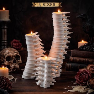 Puede incluir: Tres velas blancas con forma de columna vertebral de diferentes alturas, con llamas encendidas. Las velas están sobre una superficie de madera, con una calavera, rosas y una estatua de cuervo en el fondo. El texto "3 SIZES" está encima de las velas.