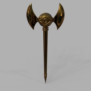 Op de afbeelding: Een gouden staf met een grote, ronde, oogvormige bol aan de bovenkant. De bol heeft een enkel oog met een zwarte pupil en een gouden iris. De staf heeft twee vleugels die zich uitstrekken vanaf de bol, waardoor een halve maan vorm ontstaat.