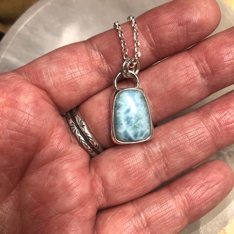 Puede incluir: Un collar de cadena de plata con un colgante que presenta una piedra preciosa azul claro y blanca engastada en un bisel de plata.