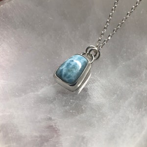 Puede incluir: Un collar de cadena de plata con un colgante de piedra preciosa azul claro. El colgante tiene una forma rectangular con una superficie lisa y pulida.