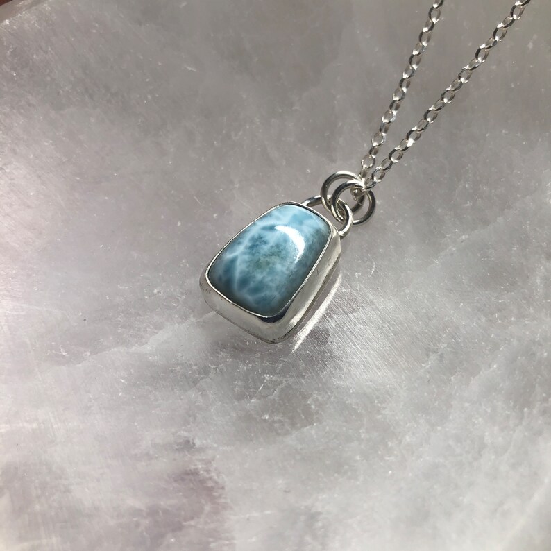 Puede incluir: Un collar de cadena de plata con un colgante de piedra preciosa azul claro. El colgante tiene una forma rectangular con una superficie lisa y pulida.