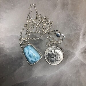 Puede incluir: Un collar de cadena de plata con un colgante de piedra preciosa azul claro. El colgante tiene forma rectangular con una superficie lisa y pulida. La piedra preciosa es de color azul claro con vetas blancas que la atraviesan. El collar mide aproximadamente 45 cm de largo y se muestra junto a una moneda de diez centavos de d&oacute;lar estadounidense para comparar el tama&ntilde;o.