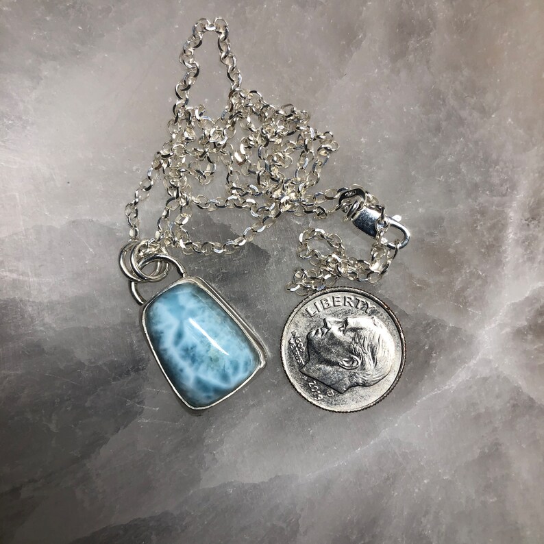 Puede incluir: Un collar de cadena de plata con un colgante de piedra preciosa azul claro. El colgante tiene forma rectangular con una superficie lisa y pulida. La piedra preciosa es de color azul claro con vetas blancas que la atraviesan. El collar mide aproximadamente 45 cm de largo y se muestra junto a una moneda de diez centavos de d&oacute;lar estadounidense para comparar el tama&ntilde;o.