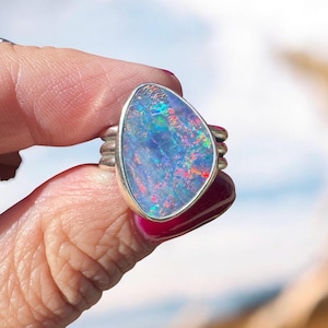 May include: A silver ring featuring a large, pear-shaped opal gemstone with a vibrant play-of-color displaying flashes of blue, green, and red. The setting consists of a smooth metal bezel surrounding the stone, mounted on a triple-band silver shank.