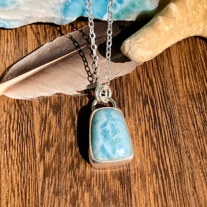 Puede incluir: Un collar de cadena de plata con un colgante de piedra preciosa azul claro. El colgante tiene forma rectangular con una superficie lisa y pulida. La piedra preciosa tiene un patr&oacute;n de remolino de azul claro y blanco.