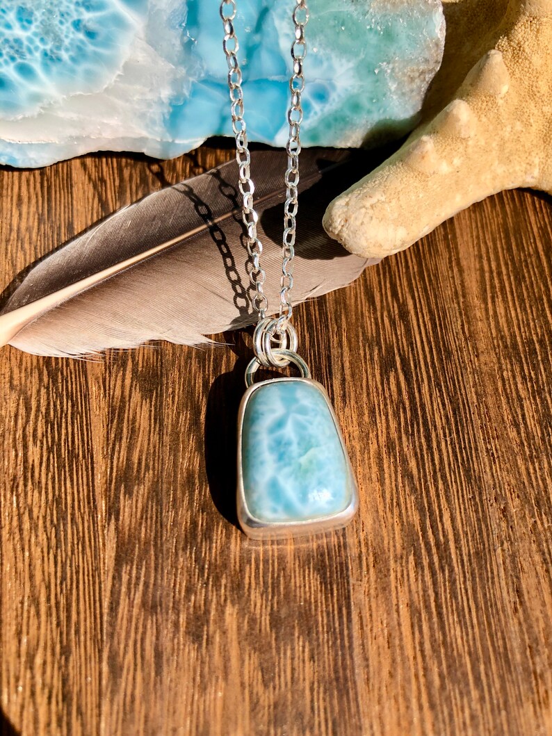 Puede incluir: Un collar de cadena de plata con un colgante de piedra preciosa azul claro. El colgante tiene forma rectangular con una superficie lisa y pulida. La piedra preciosa tiene un patr&oacute;n de remolino de azul claro y blanco.