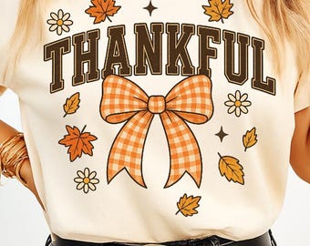 May include: A cream-coloured t-shirt with the word "THANKFUL" in brown block letters. The shirt features a plaid orange bow, autumn leaves, and floral accents. A casual, seasonal design, ideal for the fall season.