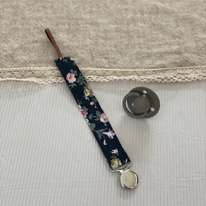 Peut inclure: Un attache-sucette à motif floral noir et blanc avec un fermoir argenté et une boucle en cuir marron. L'attache est fixée à une sucette grise.