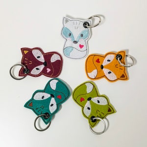 Könnte beinhalten: Fünf Schlüsselanhänger aus Filz mit gestickten Fuchs-Designs. Jeder Schlüsselanhänger zeigt einen Fuchs in einer anderen Farbe mit einem kleinen roten Herzen auf der Brust. Die Schlüsselanhänger sind an silbernen Schlüsselringen befestigt.