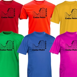 Puede incluir: Una colección de seis camisetas de manga corta en rojo, naranja, amarillo, verde, azul y rosa. Cada camiseta presenta un gráfico negro de una montaña rusa con el texto Cedar Point y el año 2026 impresos en la parte frontal del pecho.