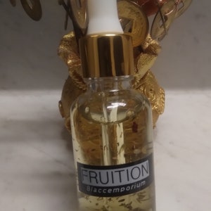 May include: A clear glass bottle with a gold lid and dropper. The bottle is filled with a light yellow liquid and contains small, gold flakes. The label on the bottle reads "FRUITION Blaccemporium".