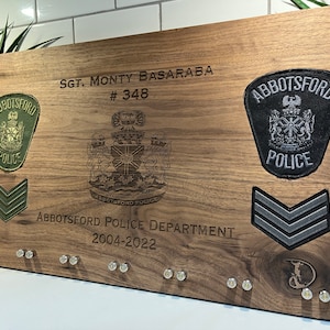 May include: A wooden plaque with a black and white engraved image of a crest, a green and black police patch, a grey and black police patch, and a grey chevron patch. The plaque also has the text "SGT. MONTY BASARABA #348 ABBOTSFORD POLICE DEPARTMENT 2004-2022".