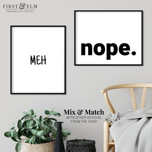 May include: Two black framed prints with white backgrounds. The print on the left says "MEH" in black text. The print on the right says "nope." in bold black text. The prints are hanging on a light gray wall.