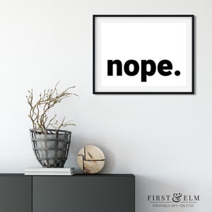 May include: Black and white printable wall art with the word "nope." in bold, black font. The print is framed in a black frame and is hanging on a white wall.