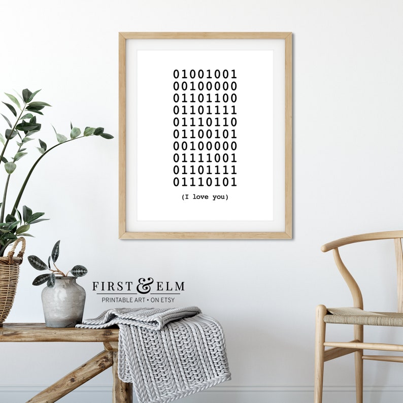 May include: A framed print with a white background and black text. The text is a series of binary code that translates to "I love you". The frame is light brown wood. The print is sitting on a wooden table with a gray blanket and a potted plant.