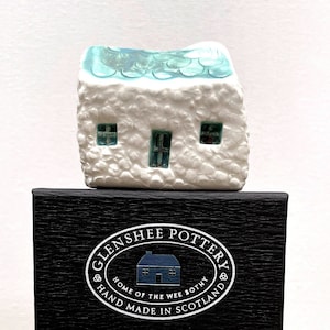 May include: A white ceramic cottage with a blue roof and windows, sitting on a black box with the text "Glenshee Pottery, Home of the Wee Bothy, Hand Made in Scotland".