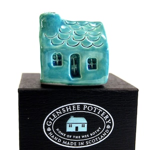 May include: A small, teal blue ceramic house with a white door and windows. The house is sitting on a black box with the words "Glenshee Pottery" and "Home of the Wee Bothy" printed on it.