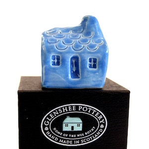 May include: A small blue ceramic house with a white roof and windows. The house is sitting on a black box with the text "Glenshee Pottery Home of the Wee Bothy Hand Made in Scotland".