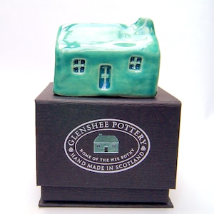 Op de afbeelding: Een klein, turquoise keramiek huisje met witte ramen en een deur. Het huisje staat op een zwarte doos met een zilveren label waarop staat "Glenshee Pottery, Home of the Wee Bothy, Hand Made in Scotland."