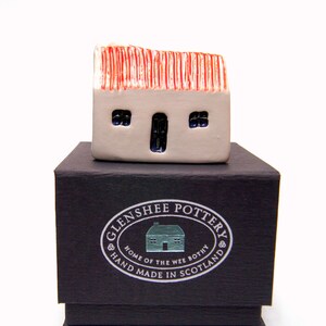 May include: A small white ceramic house with a red roof sits on top of a black box. The box has a silver label that reads "Glenshee Pottery, Home of the Wee Bothy, Hand Made in Scotland".