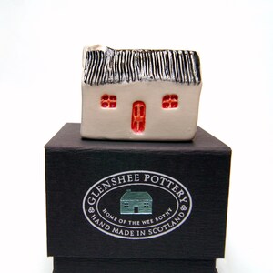 May include: A small white ceramic house with a black thatched roof and red windows. The house is sitting on a black box with a silver label that reads "Glenshee Pottery, Home of the Wee Bothy, Hand Made in Scotland."