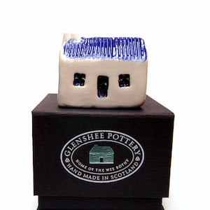 May include: A small white ceramic house with a blue roof and two windows. The house is sitting on a black box with a silver label that reads "Glenshee Pottery, Home of the Wee Bothy, Hand Made in Scotland."