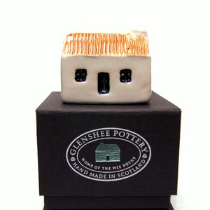 Puede incluir: Una pequeña casa de cerámica blanca con techo de paja se encuentra sobre una caja negra. La caja tiene una etiqueta plateada que dice "Glenshee Pottery, Home of the Wee Bothy, Hand Made in Scotland."