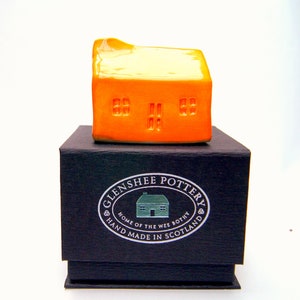 May include: A small orange ceramic house figurine in a black box with a silver label that reads "Glenshee Pottery, Home of the Wee Bothy, Hand Made in Scotland".
