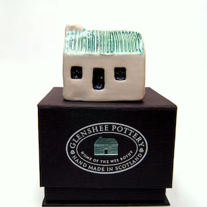 May include: A small white ceramic house with a green roof sits on a black box. The box has a silver label that reads "Glenshee Pottery, Home of the Wee Bothy, Hand Made in Scotland".