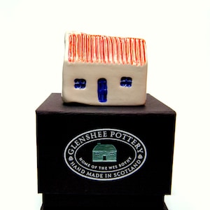 May include: A small white ceramic house with a red roof and blue windows sits on a black box. The box has a white label with the text "Glenshee Pottery, Home of the Wee Bothy, Hand Made in Scotland."