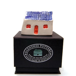 May include: A small white ceramic house with a blue roof and red door. The house is sitting on a black box with a silver label that reads "Glenshee Pottery, Home of the Wee Bothy, Hand Made in Scotland."