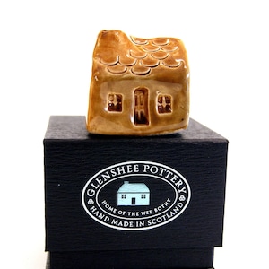 May include: A miniature ceramic house with a brown glaze, featuring a detailed roof, door, and windows. The house sits atop a black gift box with the text "Glenshee Pottery, Home of the Wee Bothy, Hand Made in Scotland."
