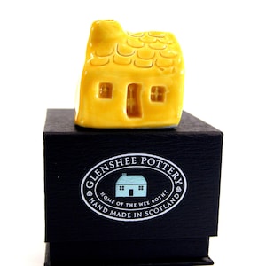 May include: A small yellow ceramic cottage with a thatched roof, sitting on a black box with a white label that reads "Glenshee Pottery, Home of the Wee Bothy, Hand Made in Scotland".