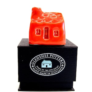 May include: A small, red ceramic house with a textured roof and three windows. The house is sitting on a black box with the words "Glenshee Pottery" and "Home of the Wee Bothy" printed on it.